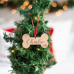 May include: A wooden bone-shaped Christmas ornament with the name "Charlie" and the year "2018" engraved on it. The ornament is hanging on a small artificial Christmas tree.