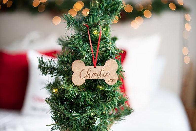 May include: A wooden bone-shaped Christmas ornament with the name "Charlie" and the year "2018" engraved on it. The ornament is hanging on a small artificial Christmas tree.