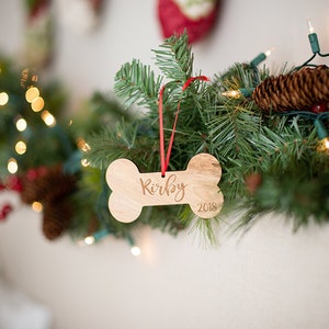 May include: A wooden dog bone ornament with the name "Kirby" and the year "2018" engraved on it. The ornament is hanging from a red ribbon on a green Christmas tree branch.