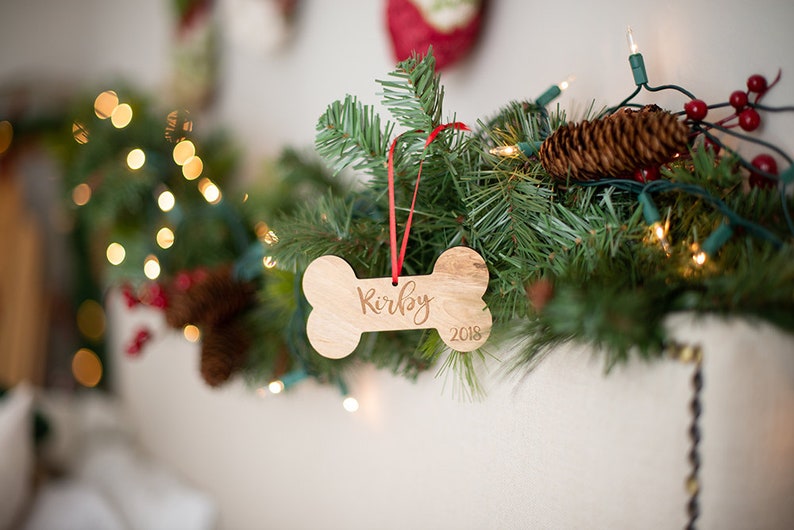 May include: A wooden dog bone ornament with the name "Kirby" and the year "2018" engraved on it. The ornament is hanging from a red ribbon on a green Christmas tree branch.