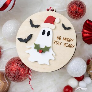 以下が含まれることがあります： サンタ帽をかぶったお化けと「Be Merry Stay Scary」の文字が入った木製のクリスマスオーナメント。オーナメントは、オーナメントやキャンディー型の装飾など、赤と白のホリデーデコレーションに囲まれています。