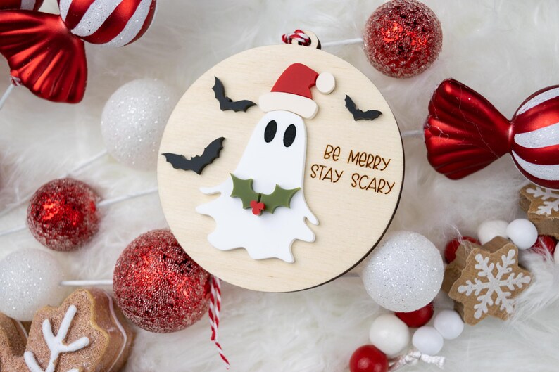 以下が含まれることがあります： サンタ帽をかぶったお化けと「Be Merry Stay Scary」の文字が入った木製のクリスマスオーナメント。オーナメントは、オーナメントやキャンディー型の装飾など、赤と白のホリデーデコレーションに囲まれています。