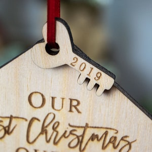 May include: A wooden Christmas ornament shaped like a house with a keyhole and a key. The key is engraved with the year 2019. The ornament is inscribed with the text "Our first Christmas in our home".