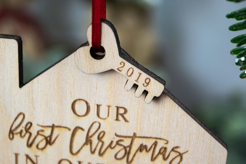 May include: A wooden Christmas ornament shaped like a house with a keyhole and a key. The key is engraved with the year 2019. The ornament is inscribed with the text "Our first Christmas in our home".