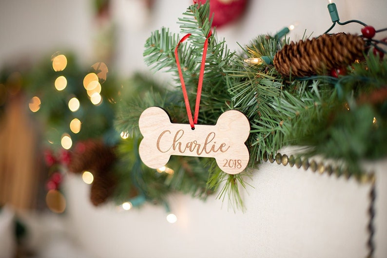 May include: A wooden dog bone ornament with the name "Charlie" and the year "2018" engraved on it. The ornament is hanging on a red ribbon from a Christmas tree.