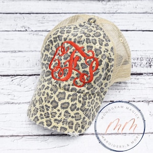 May include: A brown and beige leopard print trucker hat with a red embroidered monogram. The hat has a mesh back and a distressed look.