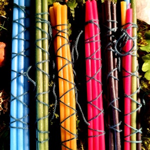 Può includere: Sei mazzi di sottili candele colorate legate con spago. Le candele sono blu, verdi, gialle, rosse, nere e bianche. I mazzi sono disposti in fila su un letto di muschio e foglie verdi.