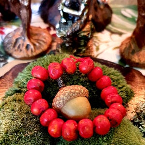 Puede incluir: Un brazalete de cuentas rojas con una bellota en el centro, descansando sobre un lecho de musgo verde.