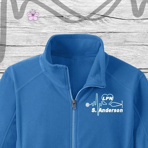May include: A royal blue fleece jacket with a white embroidered design of a stethoscope, a heart with the letters LPN inside, and a heartbeat line with the name "S. Anderson" underneath.