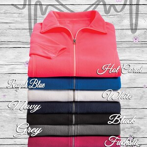 May include: A stack of fleece zip-up jackets in various colors, including hot coral, royal blue, white, navy, grey, fuchsia, and purple. The jackets are arranged from top to bottom, with the hot coral jacket on top. The text "Nurse Life" is written in a heart shape above the jackets.