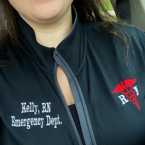 May include: Black zip-up jacket with a gray zipper and a red and white embroidered medical symbol with the letters "RN" on the right side. The text "Kelly, RN Emergency Dept." is embroidered on the left side of the jacket.