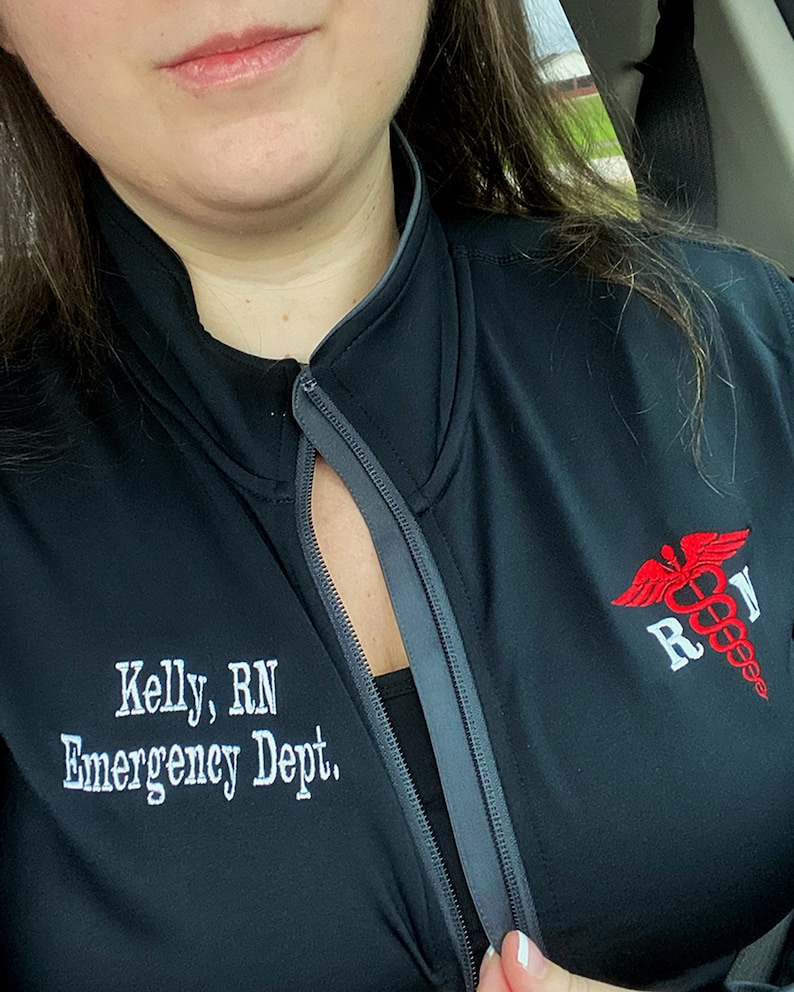May include: Black zip-up jacket with a gray zipper and a red and white embroidered medical symbol with the letters "RN" on the right side. The text "Kelly, RN Emergency Dept." is embroidered on the left side of the jacket.