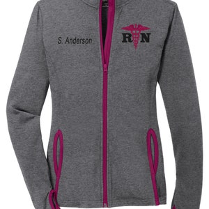 May include: A gray and pink zip-up jacket with a pink zipper and pink accents. The jacket has a small embroidered logo on the right chest that says "RN" with a medical symbol. The name "S. Anderson" is embroidered on the left chest.