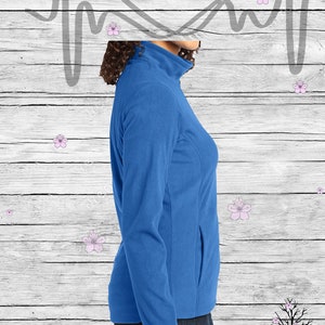 May include: A royal blue fleece jacket with a zipper closure and a single pocket on the left side. The jacket is worn by a person with brown hair and is shown from the side. The background is a white woodgrain pattern with pink flowers.
