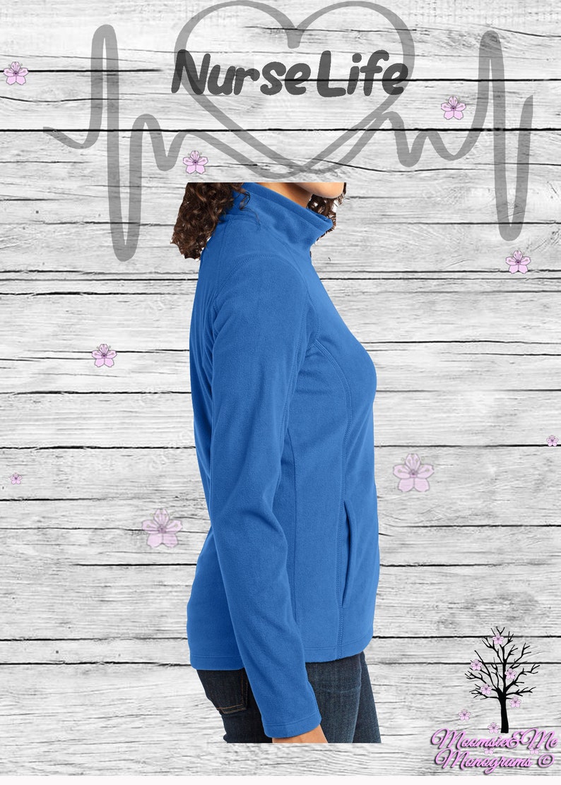 May include: A royal blue fleece jacket with a zipper closure and a single pocket on the left side. The jacket is worn by a person with brown hair and is shown from the side. The background is a white woodgrain pattern with pink flowers.