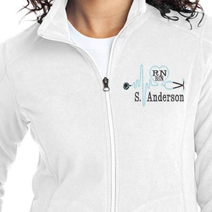 May include: White zippered jacket with a blue and black embroidered design of a heart with the text "RN BSN" and a stethoscope with a heartbeat line. The text "S. Anderson" is below the design.