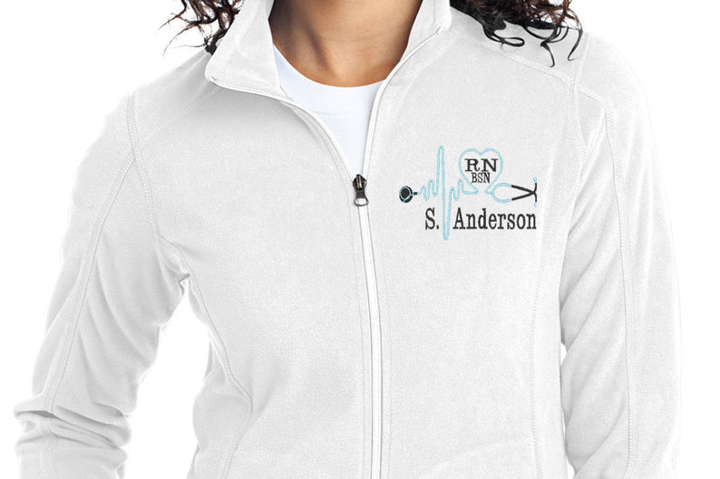 May include: White zippered jacket with a blue and black embroidered design of a heart with the text "RN BSN" and a stethoscope with a heartbeat line. The text "S. Anderson" is below the design.