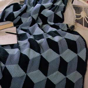 May include: A blue and navy blue crocheted blanket with a 3D geometric pattern. The blanket is folded and draped over a white bed.