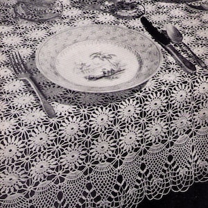 May include: A white crocheted tablecloth with a delicate floral pattern. The tablecloth features a scalloped edge and is laid out on a dark surface. A white plate with a floral design is placed on the tablecloth, along with a fork and knife.