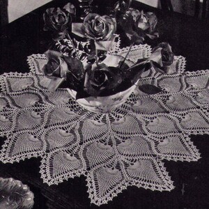 May include: A white crocheted table runner with a scalloped edge and a delicate floral pattern. The runner is laid on a dark wooden table with a bouquet of roses in a white vase.