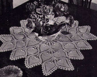 May include: A white crocheted table runner with a scalloped edge and a delicate floral pattern. The runner is laid on a dark wooden table with a bouquet of roses in a white vase.