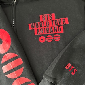 May include: A black zip-up hoodie featuring red screen-printed text that reads BTS WORLD TOUR ARIRANG with three circular symbols below. The sleeve displays large red circular graphics and the text BTS is printed on the cuff area.