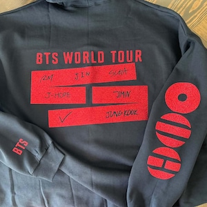 May include: A black long-sleeve hoodie featuring red text and graphics. The back displays "BTS WORLD TOUR" above red rectangular blocks containing the names RM, JIN, SUGA, J-HOPE, JIMIN, and JUNG KOOK. The sleeve features the BTS logo and text.