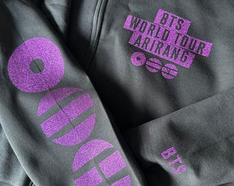 May include: A black zip-up hoodie featuring purple glitter text and graphics. The chest displays 'BTS WORLD TOUR ARIRANG' with three circular icons. The sleeve features a vertical sequence of four large circular and semi-circular purple glitter shapes.