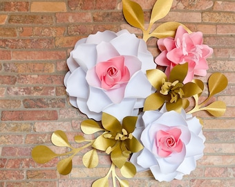 Puede incluir: Un arreglo de flores de papel con flores blancas y rosas y hojas doradas. Las flores están superpuestas y tienen un efecto 3D. Las hojas también están superpuestas y tienen un acabado dorado metálico.