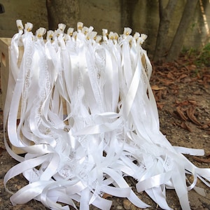 May include: White wedding wands with satin ribbons and lace accents. Each wand features a gold bell and is tied with a satin ribbon. The wands are arranged in a wooden crate, perfect for a wedding ceremony.