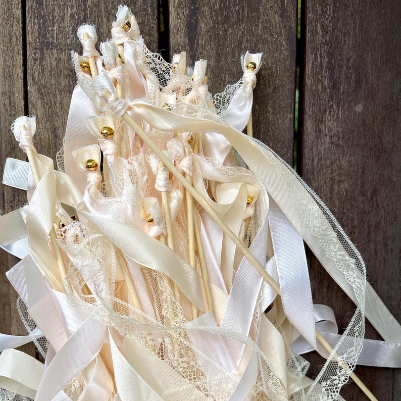 May include: A collection of ivory-colored wedding wands with wooden dowels, adorned with ribbons, lace, and gold accents. The wands feature various textures and patterns, creating an elegant and festive look. Perfect for a wedding or special event.
