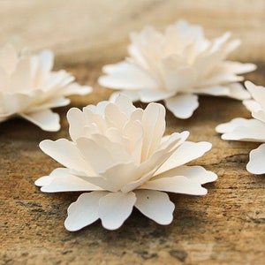 May include: Four white paper flowers with layered petals, arranged on a brown wooden surface.