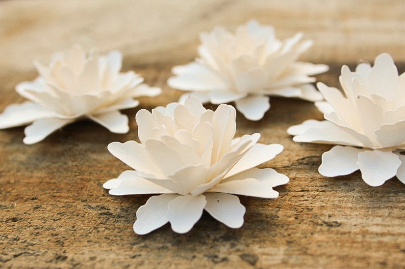 May include: Four white paper flowers with layered petals, arranged on a brown wooden surface.