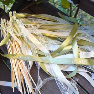 Op de afbeelding: Bruiloftstokken met groene en witte linten, versierd met delicate kant. De stokken hebben houten stokjes. De linten zijn voorzien van de namen "Alice & Edward" en de datum "11 oktober 2025".