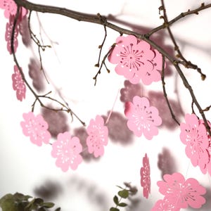 Könnte beinhalten: Girlande aus rosa Papierblumen, die an einem Ast hängen. Die Blumen haben ein filigranes, ausgeschnittenes Design und sind mit dünnen weißen Schnüren verbunden. Der Hintergrund ist weiß, mit Schatten.