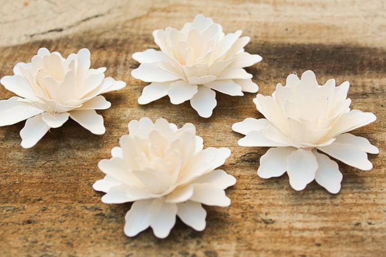 May include: Four white paper flowers with layered petals, arranged on a wooden surface.