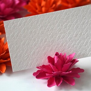 May include: White card with a textured surface, standing upright on a pink paper flower. The card is blank and ready for a message. The flower is surrounded by other paper flowers in shades of pink and orange.