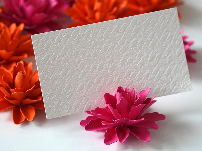 May include: White card with a textured surface, standing upright on a pink paper flower. The card is blank and ready for a message. The flower is surrounded by other paper flowers in shades of pink and orange.