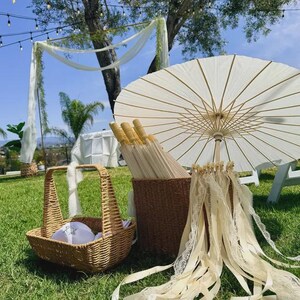 May include: A wedding scene with a white parasol, a basket, and a woven basket filled with parasols. The parasols have cream-colored paper canopies and wooden handles. The scene is set outdoors on a grassy lawn.