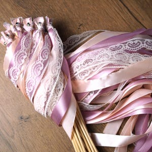 May include: A collection of wedding wands with ribbons and lace in shades of pink and lavender. The wands have wooden dowels and are adorned with satin ribbons and delicate lace trim, perfect for a wedding or special event.