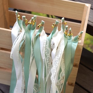 Könnte beinhalten: Hochzeitsstäbe mit weißen, mintgrünen Bändern und Spitze, jeweils verziert mit goldenen Glöckchen und Bindfaden. Die Stäbe sind in einer Holzkiste angeordnet. Ein rustikales Hochzeitsdekor.