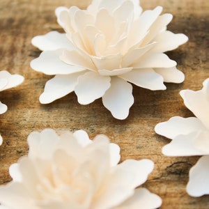 May include: Four white paper flowers with a layered, ruffled design. The flowers are arranged on a wooden surface.