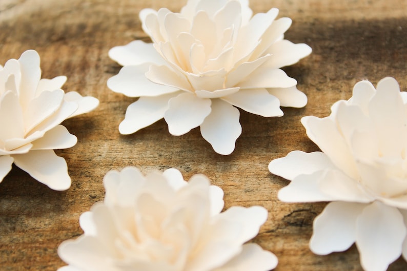 May include: Four white paper flowers with a layered, ruffled design. The flowers are arranged on a wooden surface.