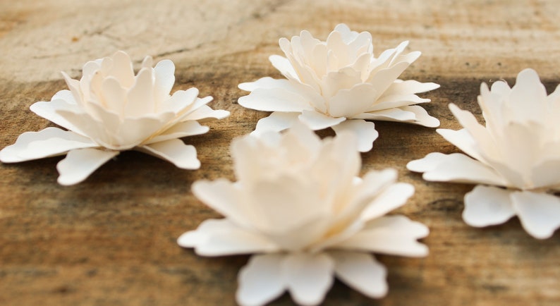 May include: Four white paper flowers with layered petals, arranged on a wooden surface.