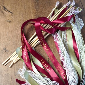 Peut inclure: Des baguettes de mariage avec des rubans bordeaux, de la dentelle et des bâtons en bois. Les rubans portent les noms "Alejandro & Ariana" et la date "12.12.26". Les baguettes sont disposées sur une surface en bois.