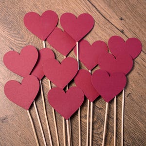May include: Burgundy paper heart decorations on wooden sticks. The hearts are arranged on a wooden surface. The image shows a collection of heart-shaped decorations, perfect for Valentine's Day or other romantic occasions.