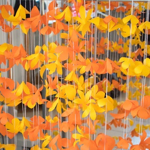 Può includere: Una decorazione da parete realizzata con fiori di carta arancioni e gialli appesi a delle corde. I fiori sono disposti in file e sono tutti della stessa dimensione e forma.