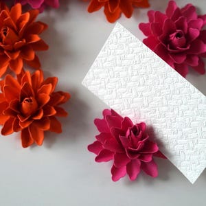 May include: A white card with a textured surface is surrounded by orange and pink paper flowers. The flowers are arranged in a circle around the card.