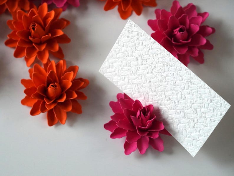 May include: A white card with a textured surface is surrounded by orange and pink paper flowers. The flowers are arranged in a circle around the card.