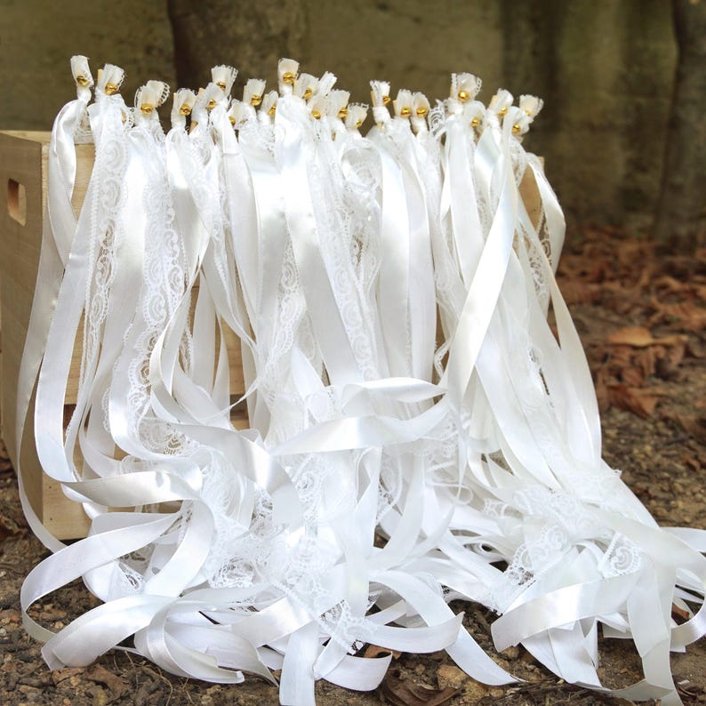 May include: A collection of white ribbons, some with delicate lace detailing, are arranged in a wooden crate. Each ribbon is adorned with a small gold bead at the top. The ribbons are of varying lengths, creating a layered effect. The background is a natural outdoor setting.